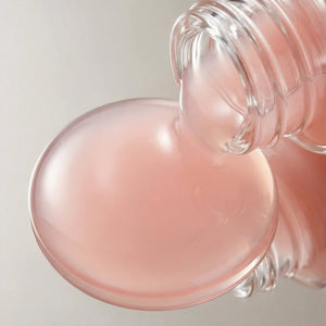 Close-up of a pink serum pouring our of a transparent bottle on a light gray background.