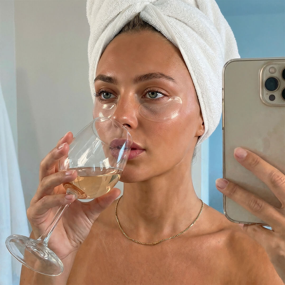 Woman with a towel on her head drinking out of a glass and using a phone, with a blue wall in the background.